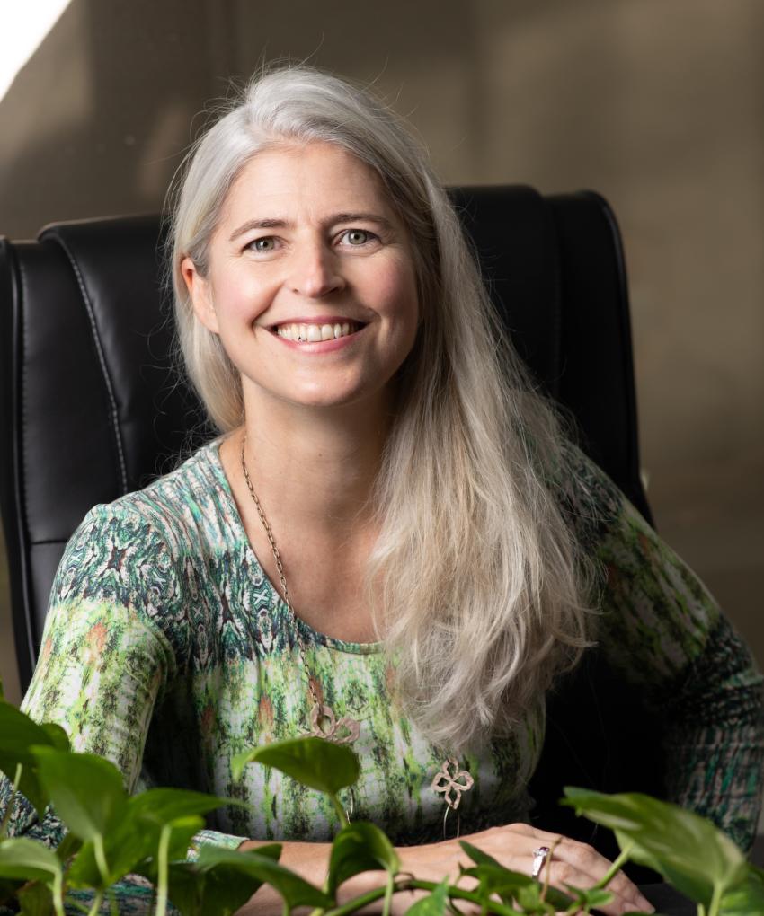 A woman with long grey hair smiles while seated at a desk indoors. She wears a patterned green top and sits in a black chair, with leafy green plants in the foreground and soft natural light illuminating the scene.