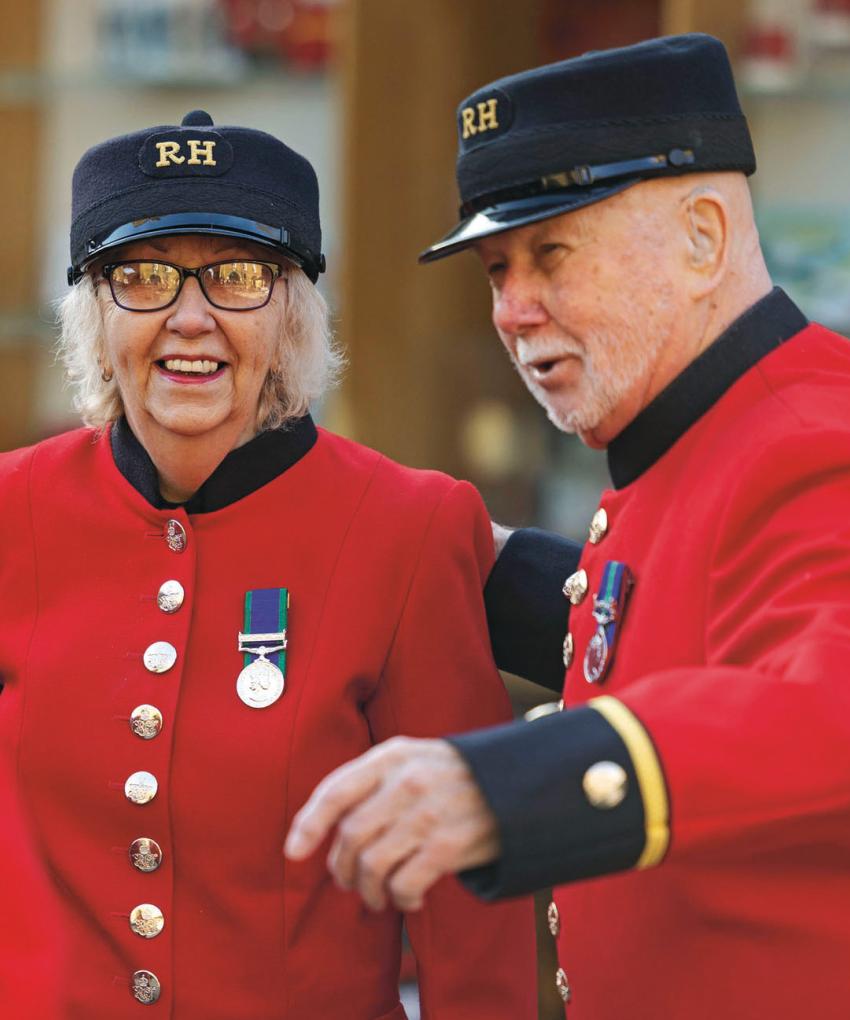 A group of Chelsea Pensioners stand together in their iconic Scarlet uniform and black Shako hat. They are laughing and smiling.