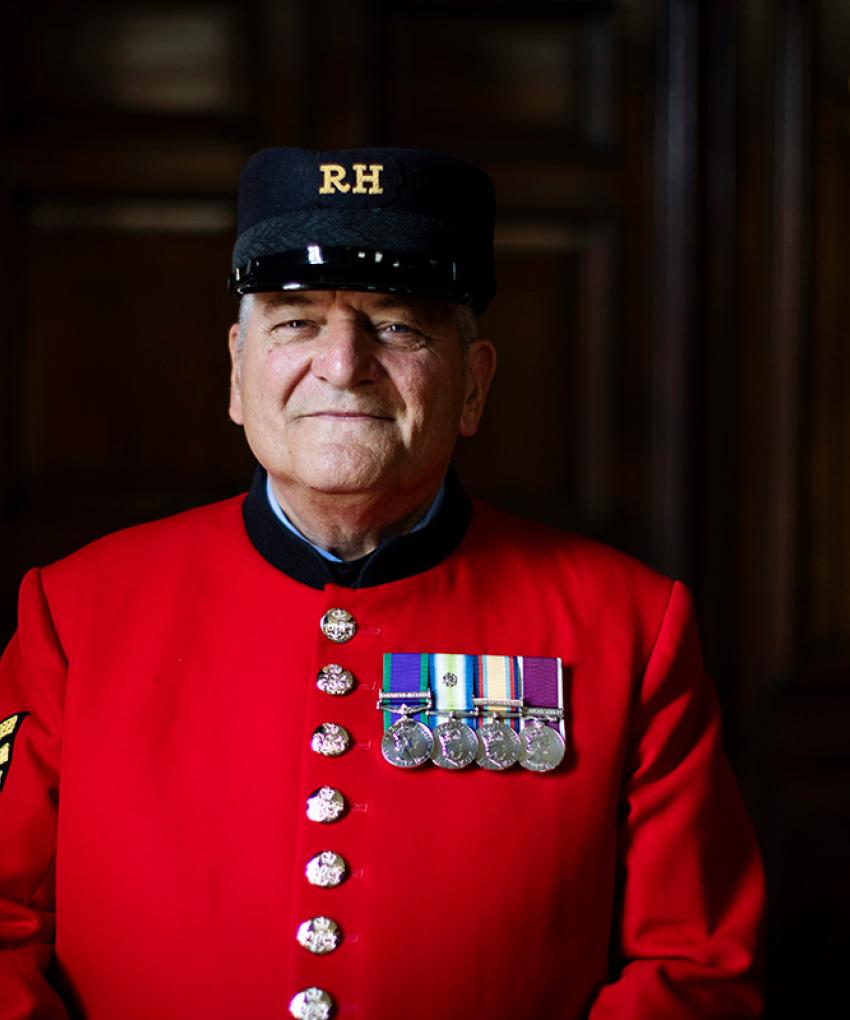 A Chelsea Pensioner stands in a wooden panelled room with his iconic scarlet uniform and a chest full of medals.