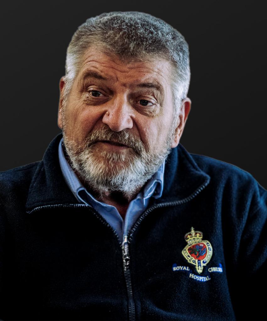 A Chelsea Pensioner sits with his blue day uniform and jacket emblazoned with The Royal Hospital Chelsea crest, set against a dark black background