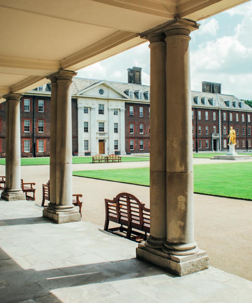 View across Figure Court at the Royal Hospital Chelsea from beneath a covered colonnade, with historic red brick buildings, manicured lawns and the statue of King Charles II visible in the distance.