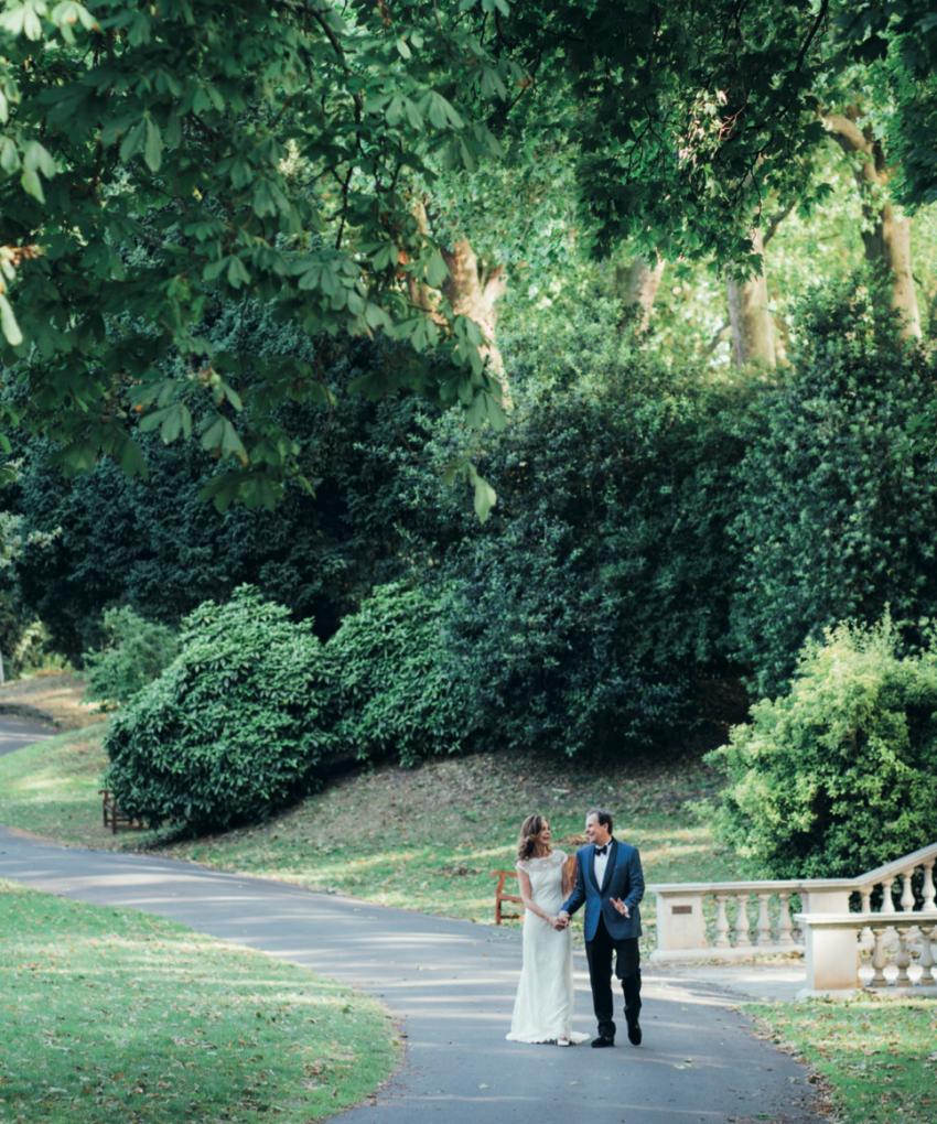 A couple walk hand in hand along a tree-lined path in the Ranelagh Gardens at the Royal Hospital Chelsea, with mature woodland planting and the historic stone staircase in the background.