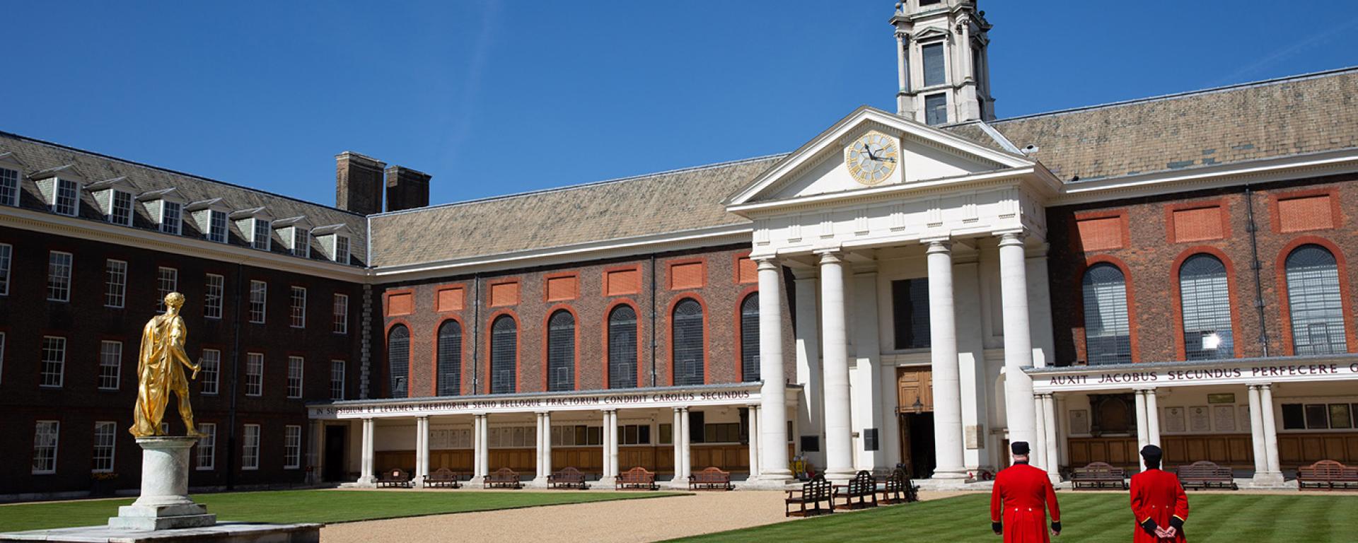 Chelsea Pensioners standing with their back to camera in Figure Court looking towards The Royal Hospital Chelsea. Gold statue of King Charles II in the distance