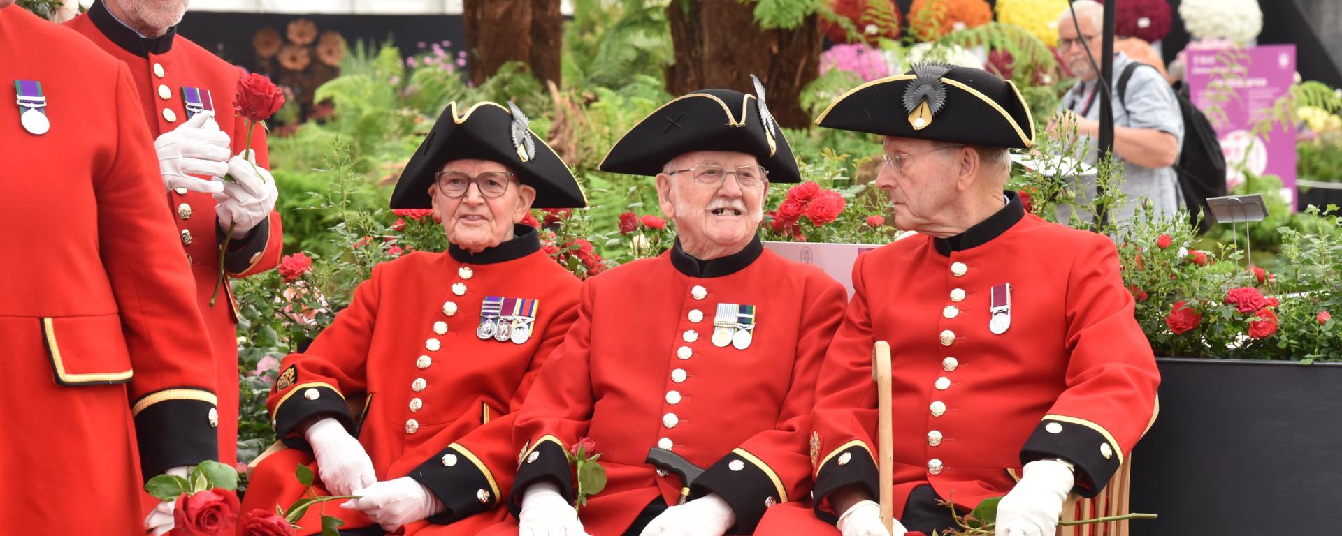 Three Chelsea Pensioners in Scarlet coats sat on a bench surrounded by flowers and plants