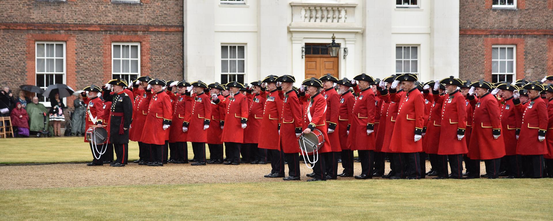 Dozens of Chelsea Pensioners stand on parade in their iconic scarlet uniforms and raise a hand in salute during the Founders Day 2025 parade.