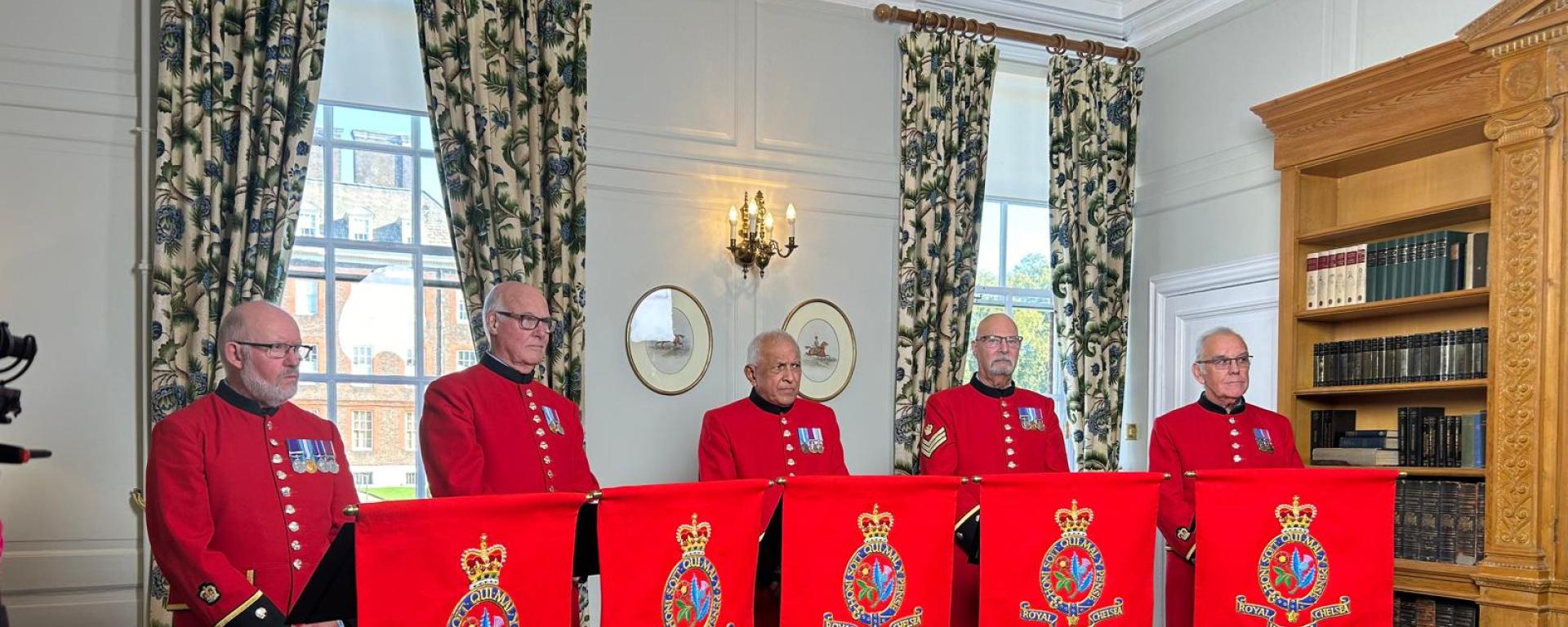 A group of Chelsea Pensioner Singers stand behind music stands in their iconic scarlet uniforms inside a room adorned with long curtains and integrated wooden bookcases