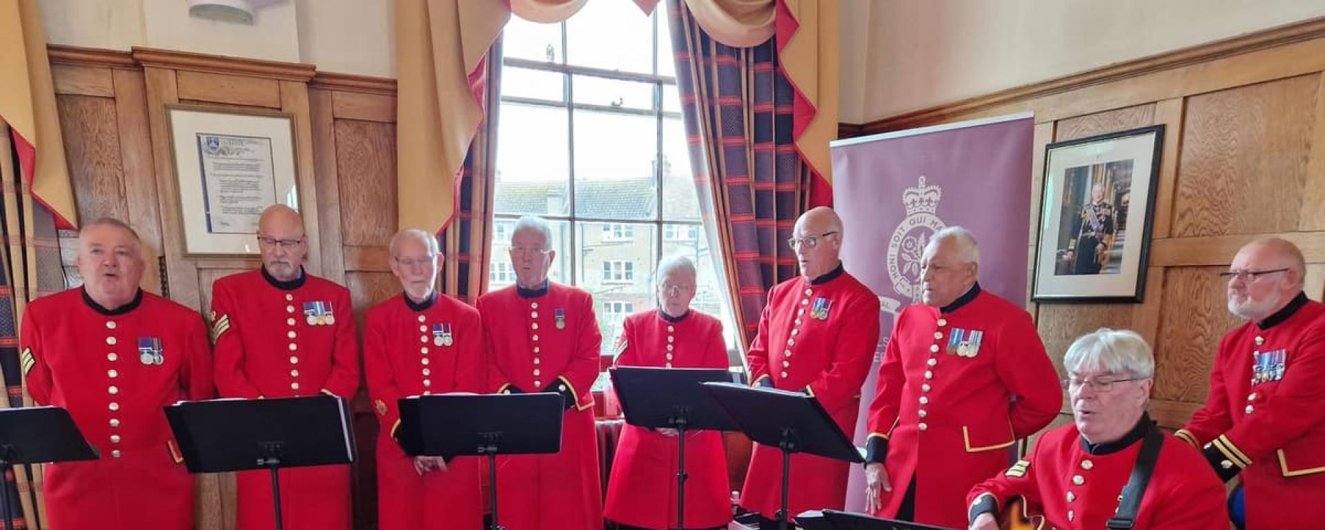 A group of scarlet clad Chelsea Pensioners are arranged behind a series of music stands in a wood panelled room with dark red carpet