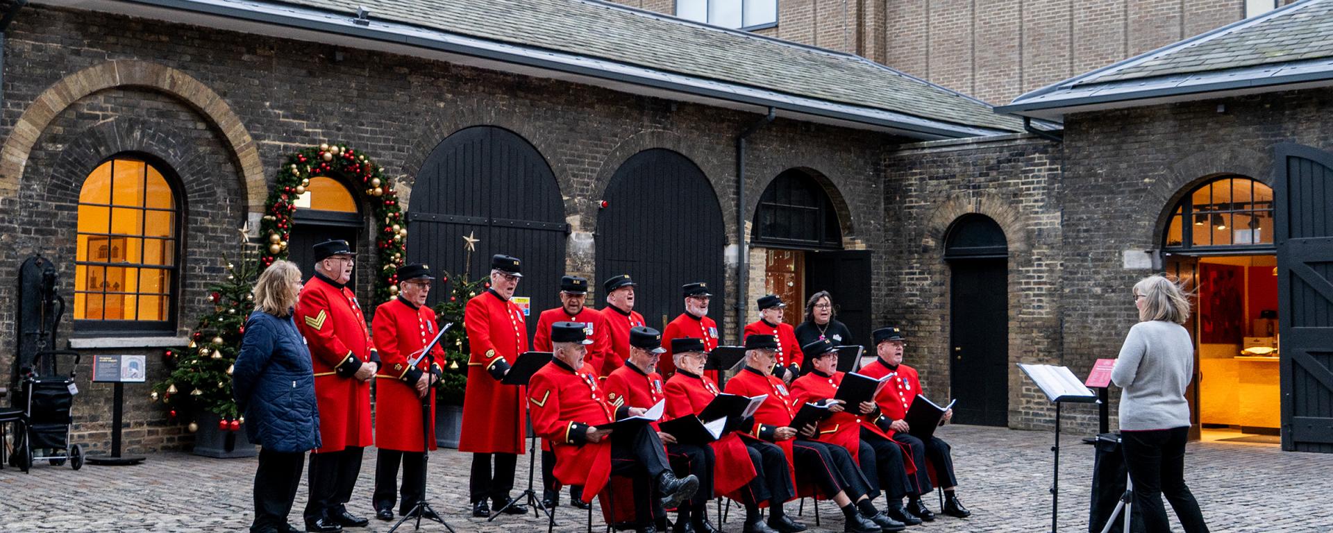 Two rows of Chelsea Pensioners, the front row seated and back row standing, wear their iconic scarlet uniforms as they sign in a stone courtyard, decorated with green, red and gold Christmas trees and garlands