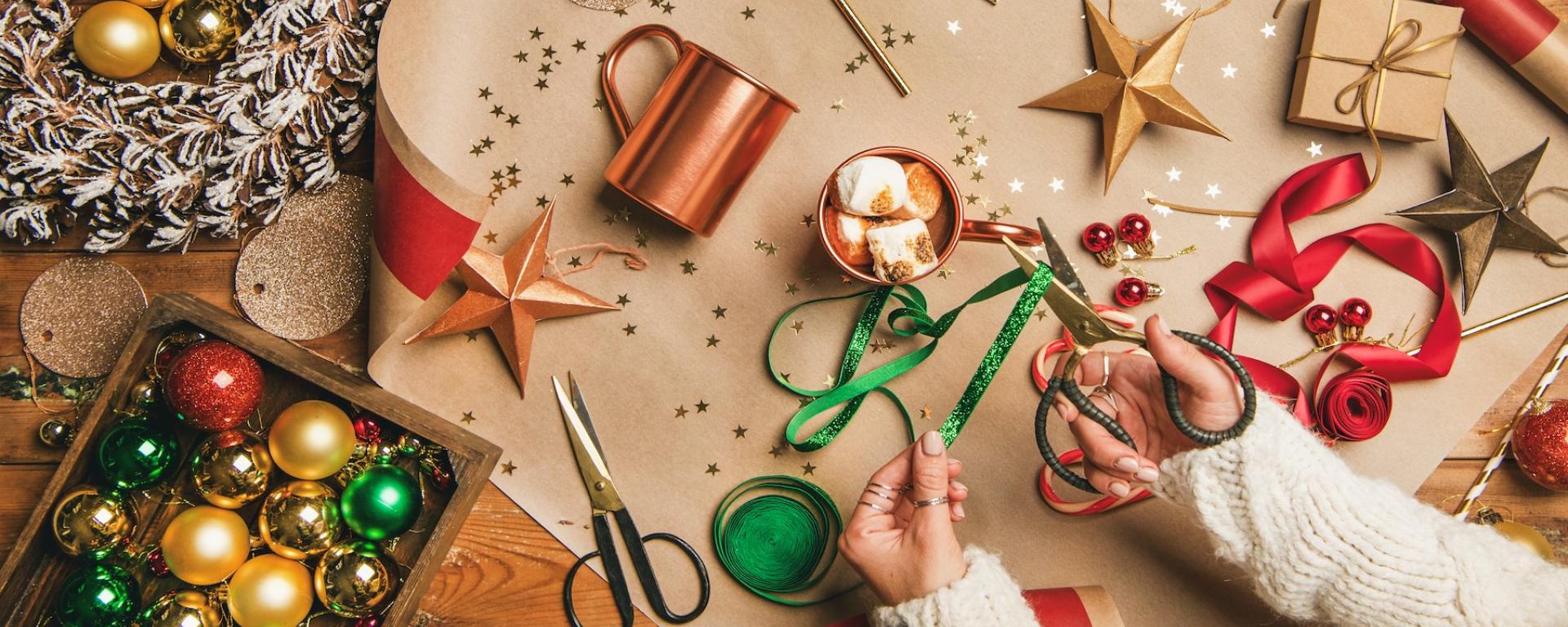 A colourful table top is shown from above with red, yellow and green decorations and crafting equipment. A pair of hands are crafting decorations while a mug of hot chocolate with huge fluffy white marshmallows sits on the table.