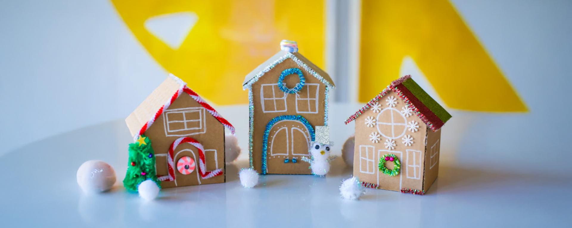 A trio of decorated cardboard gingerbread houses sit atop a white table top with a yellow backdrop on the wall behind.