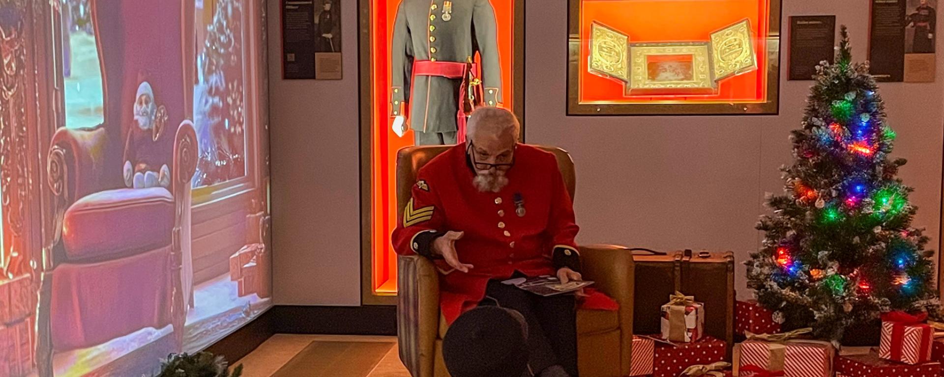 A Chelsea Pensioners sits in front of a young child in a Christmas themed grotto. A Christmas tree and lights twinkle in the corner and a digital backdrop extends the theme.