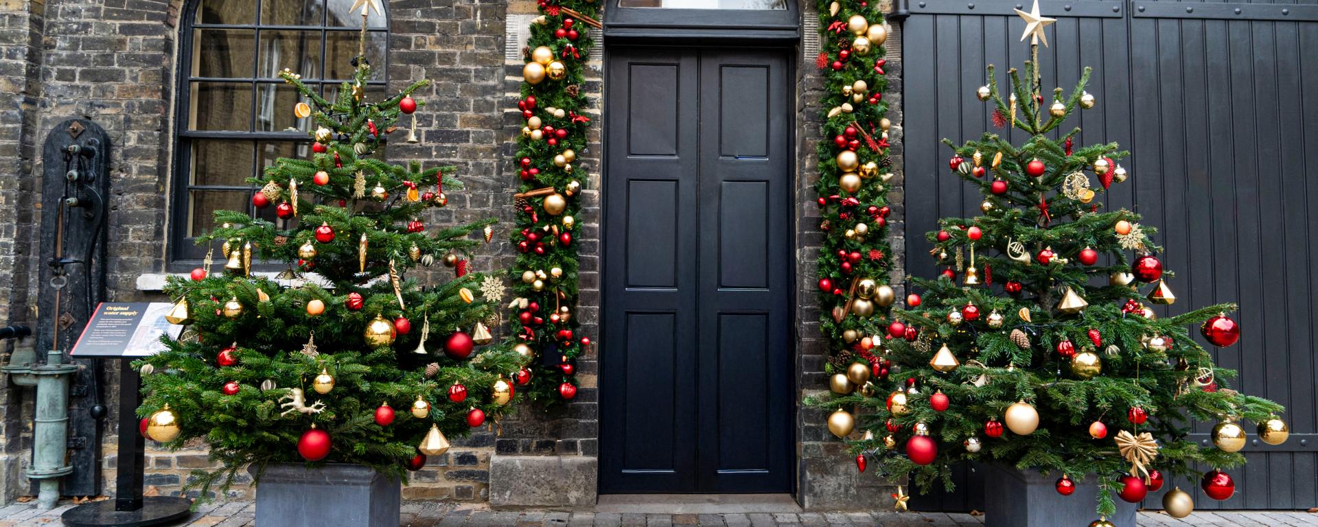 Two beautifully decorated gold, silver and red Christmas Trees stand in an outdoor courtyard besides a big black archway door with a festival garland surrounding. 