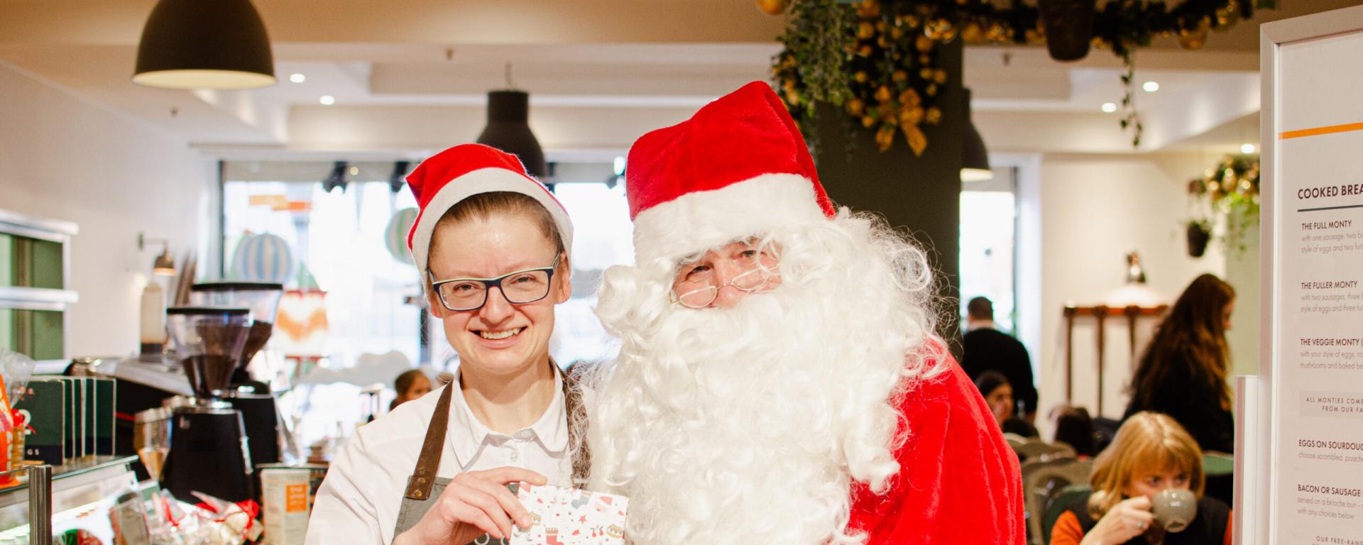 Santa is pictured in his traditional costume alongside a coffee shop employee in a red Santa hat. Indoors within a cafe setting with a counter on the left, large menu on the right and Christmas decorations in the background.