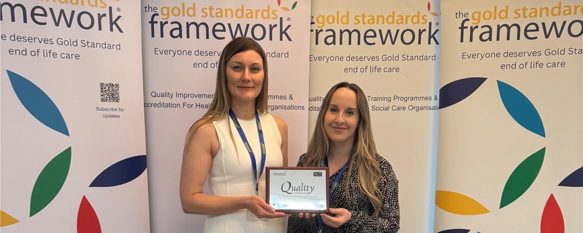 Royal Hospital Chelsea's Matron dressed in a long while dress, and Deputy Matron in a dark pattern dress, stand in front of a coloured Gold Standards Framework banner holding an award for Outstanding End of Life Care