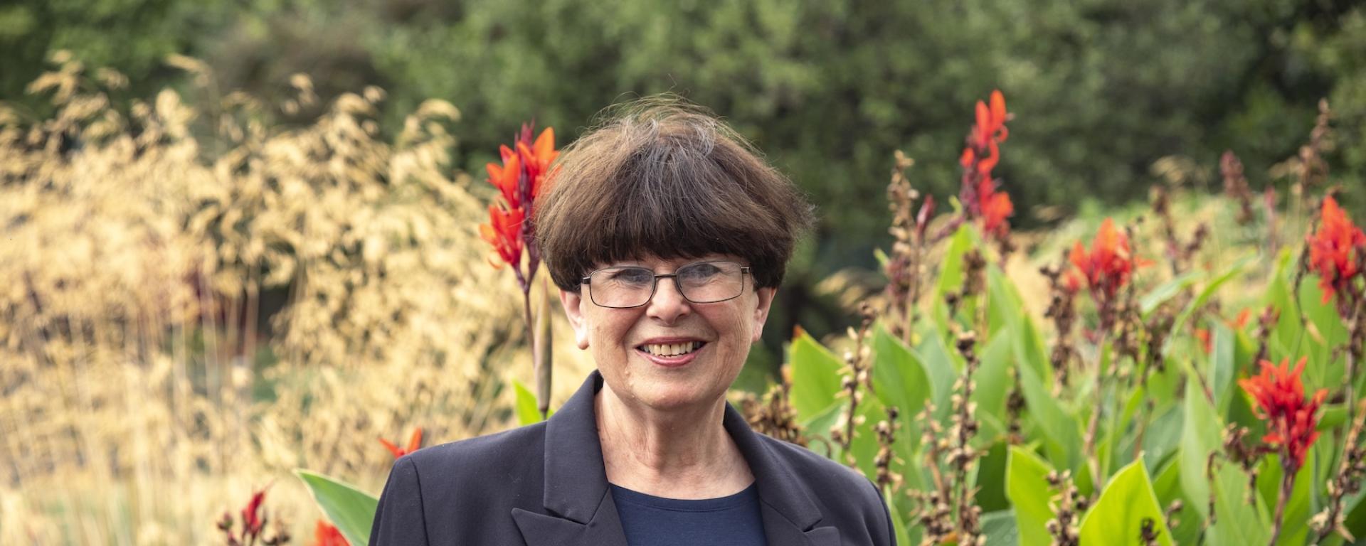 A lady with short dark hair and wearing glasses, stands in a wild garden with heavy foliage and red flowers. In contrast she's wearing a dark navy blazer and dark blue top with a Chelsea Pensioner lapel badge pinned to her jacket.