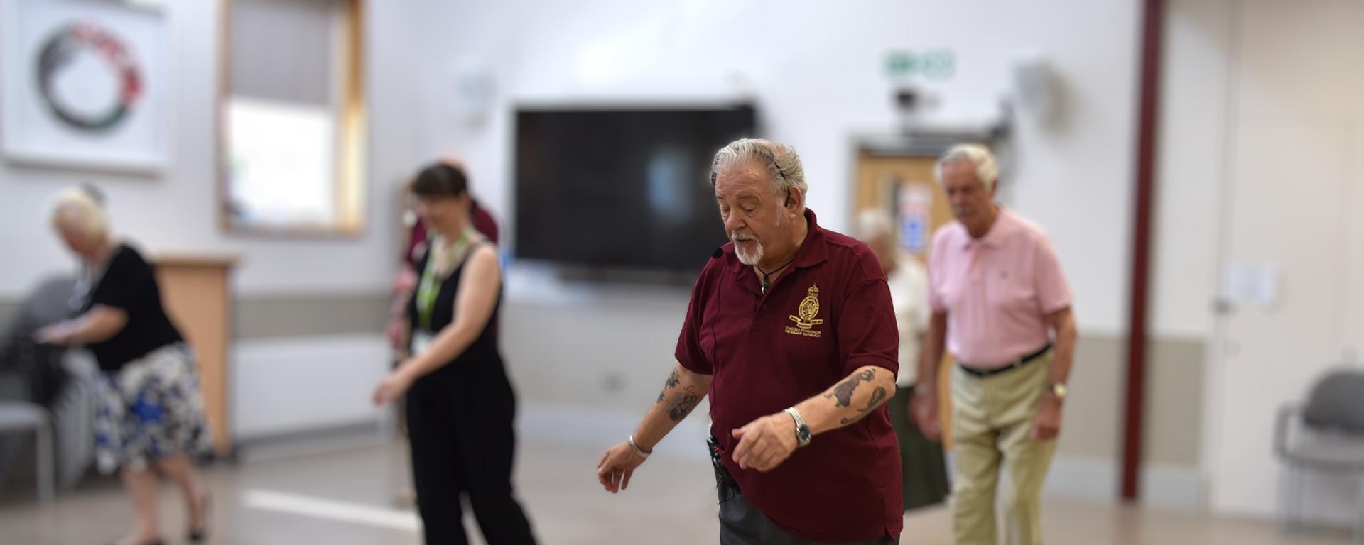 A retired older man stands at the front of a class of other adults, with a headset, instructing them in dancing.