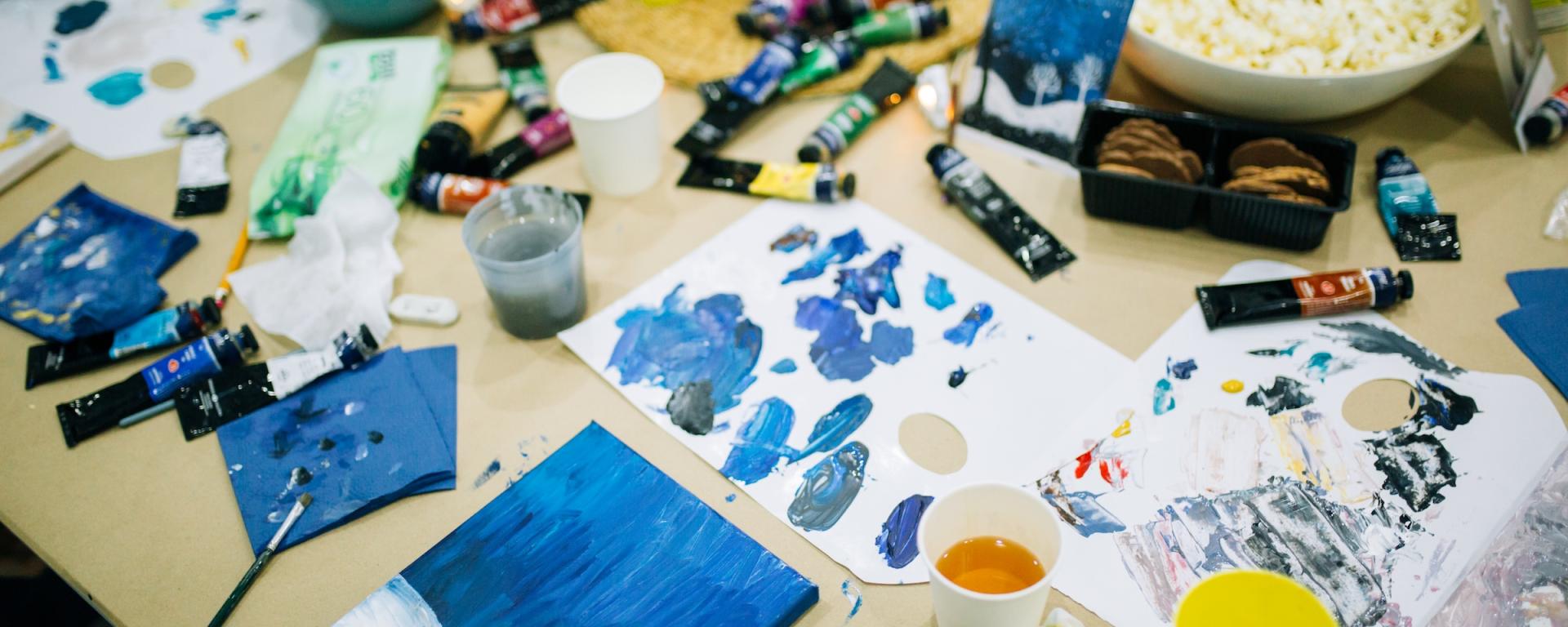 A table scattered with art suppliers, including paints, canvases, paintbrushes. Blue colours dominate, a glass of yellow liquid and a bowl of popcorn also sit upon the table.