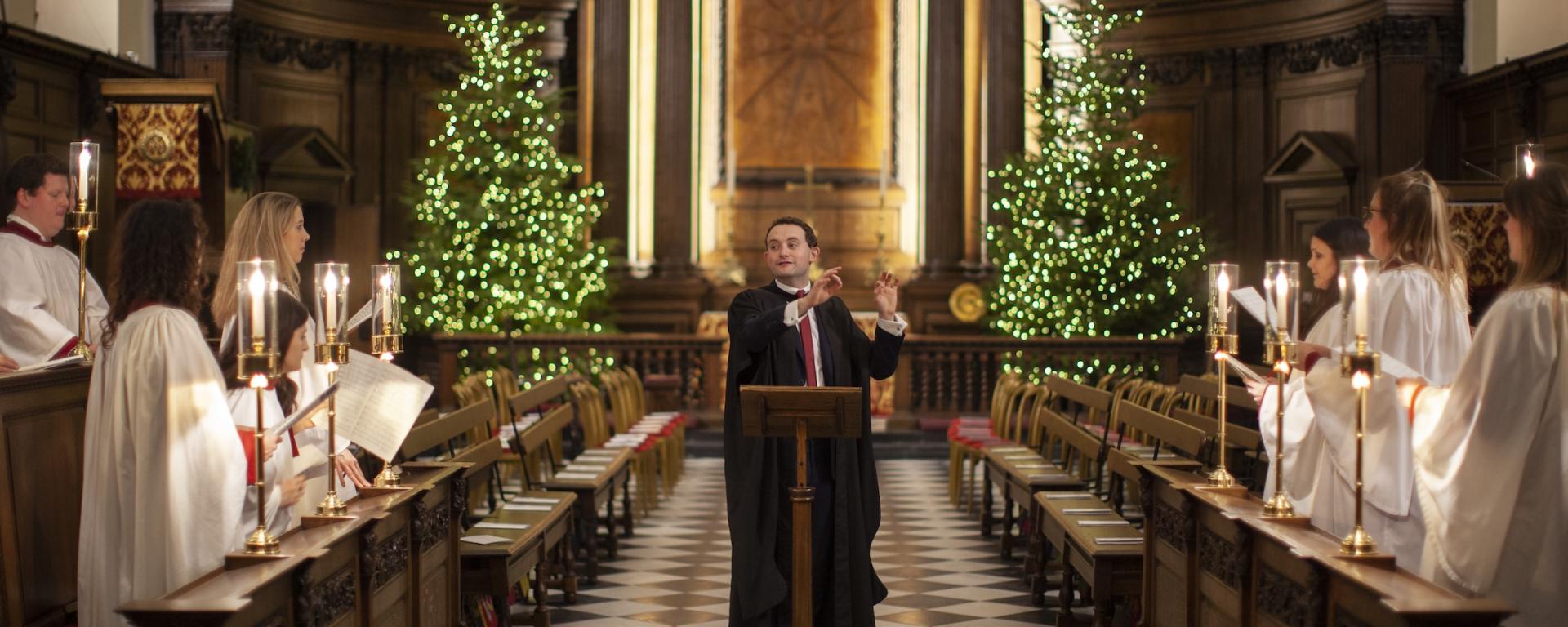 A choir performs inside a historic chapel decorated for Christmas. A conductor stands at a wooden lectern in the centre of a black and white chequered aisle, directing singers dressed in white robes who stand on either side holding candles and sheet music. Two illuminated Christmas trees flank the ornate wooden altar at the back of the chapel, creating a warm and festive atmosphere.