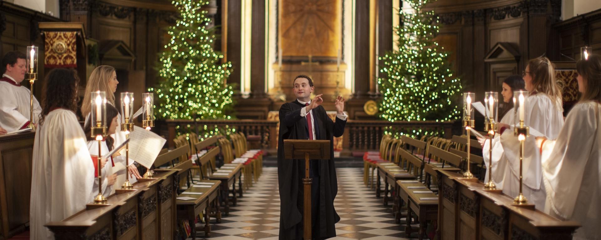 A choir performs inside a historic chapel decorated for Christmas. A conductor stands at a wooden lectern in the centre of a black and white chequered aisle, directing singers dressed in white robes who stand on either side holding candles and sheet music. Two illuminated Christmas trees flank the ornate wooden altar at the back of the chapel, creating a warm and festive atmosphere.
