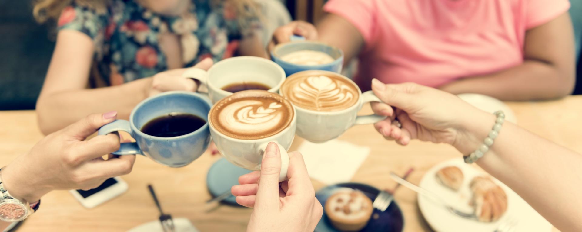 A group of people hold various mugs and cups full of tea and coffee in a collective "cheers" - beneath the cups is a wooden table with saucers, spoons and various pastries strewn about it.