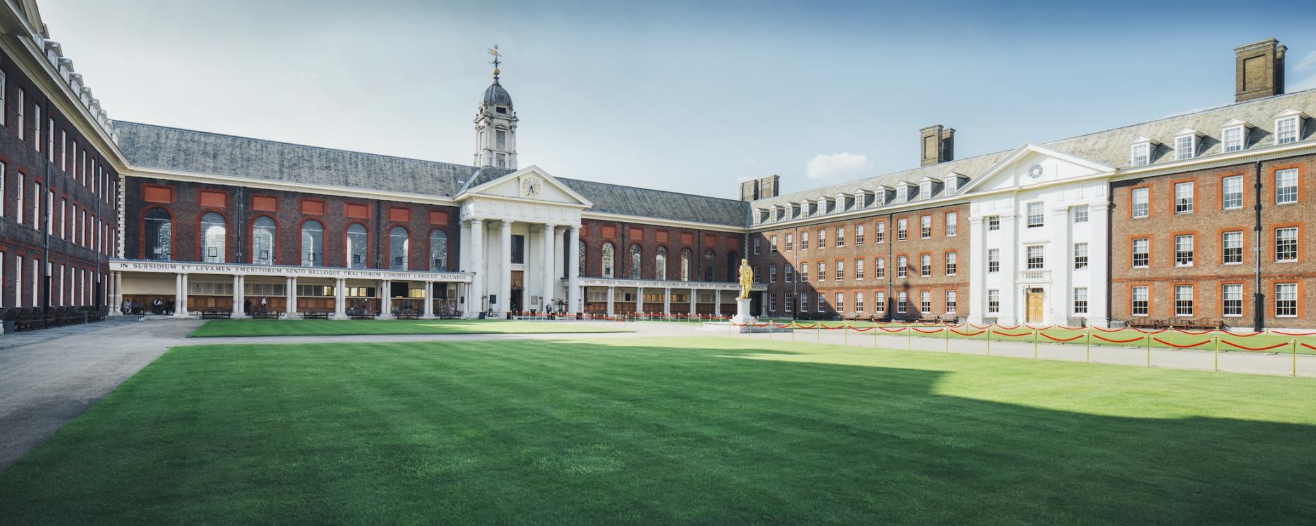 A wide view of the Main Court at the Royal Hospital Chelsea, showing an expansive green lawn surrounded by red brick buildings with white stone detailing. The central building features large columns, a pediment with a clock, and a bell tower above. A golden statue stands on a plinth to the right of centre. The scene is bright and clear, highlighting the symmetry and historic architecture of the courtyard.