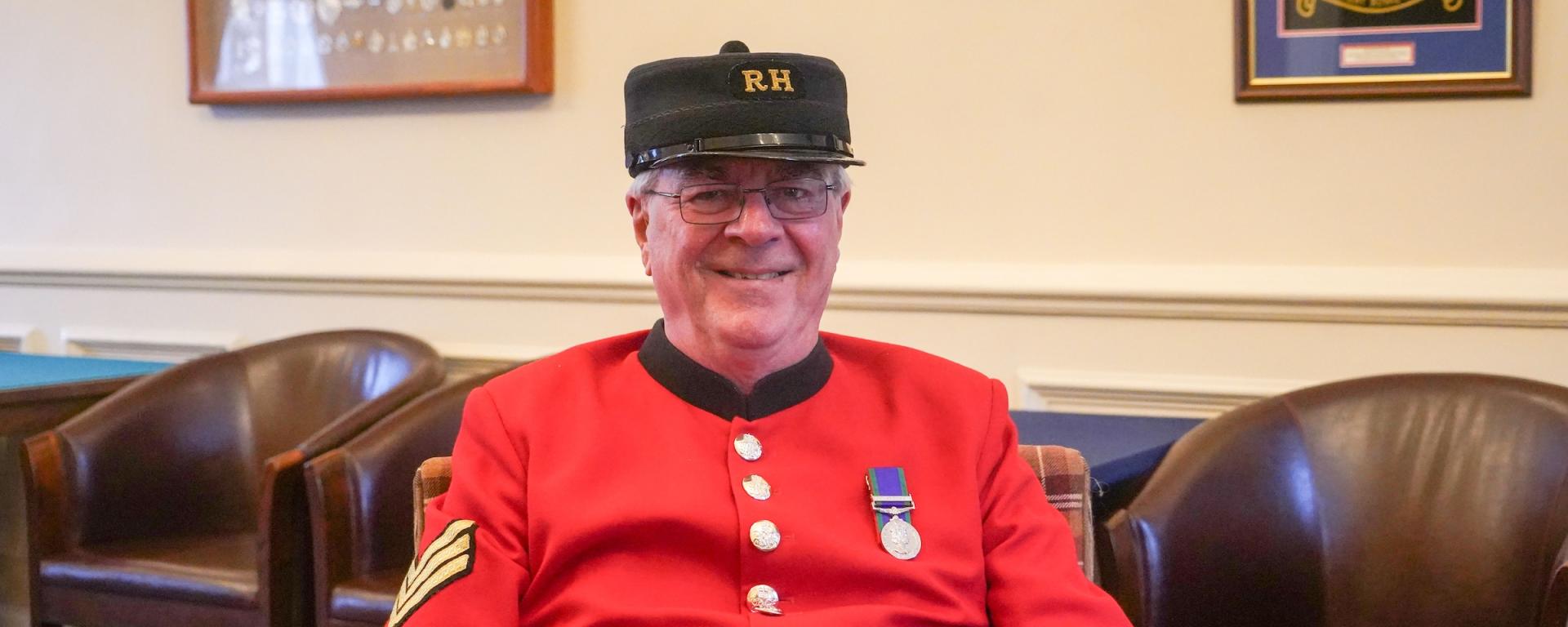 A male Chelsea Pensioner sits on a brown armchair, dressed in iconic scarlet uniform and black shako hat. A single medal adorn's his left breast.