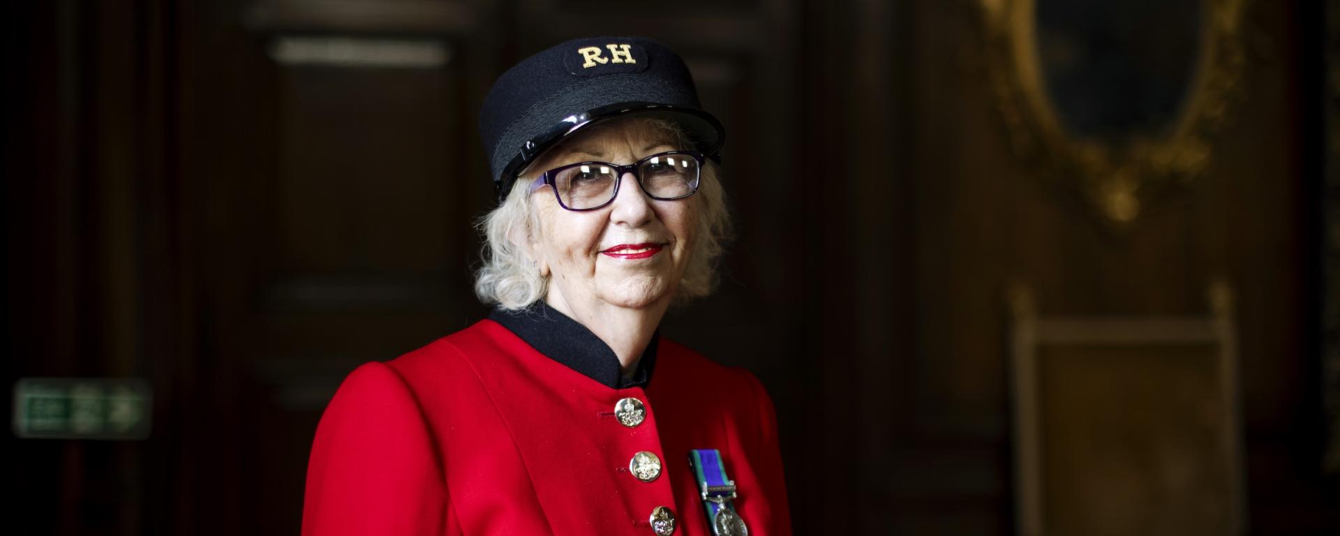 A female Chelsea Pensioner stands wearing her vibrant scarlet uniform, black shako hat and glasses, in front of large wood panelled doors.