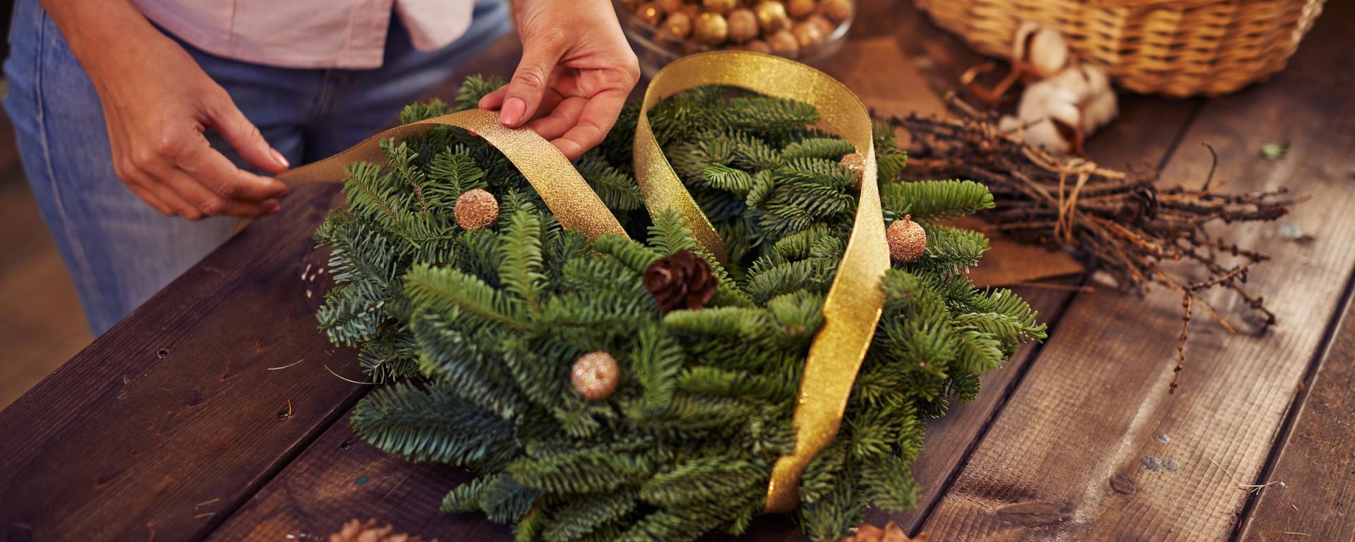 A pair of hands are tying a yellow ribbon on to a handcrafted Christmas wreath of green pine branches. A wooden table holds a container of gold baubles and pine cones.