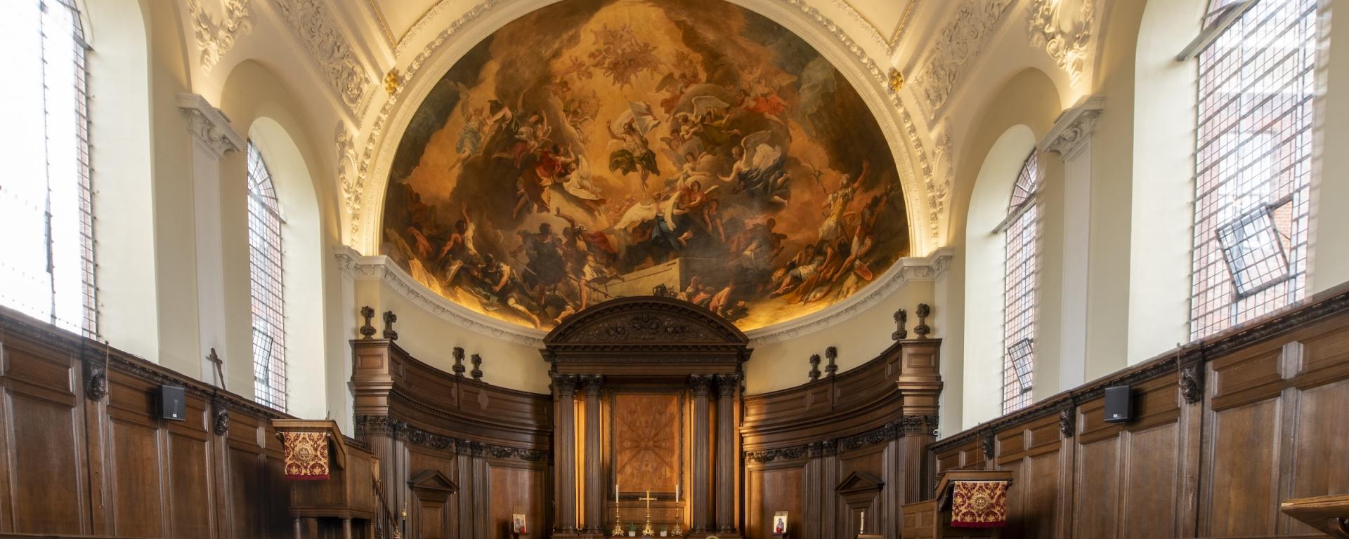 The Wren Chapel at The Royal Hospital Chelsea. Featuring wooden pews and benches on either side of a central aisle. Floor is tiled in black and white chequers and a grand alter standards at the far end. Wood panelling throughout and a huge ornate mural in a dome shape sits atop the altar.
