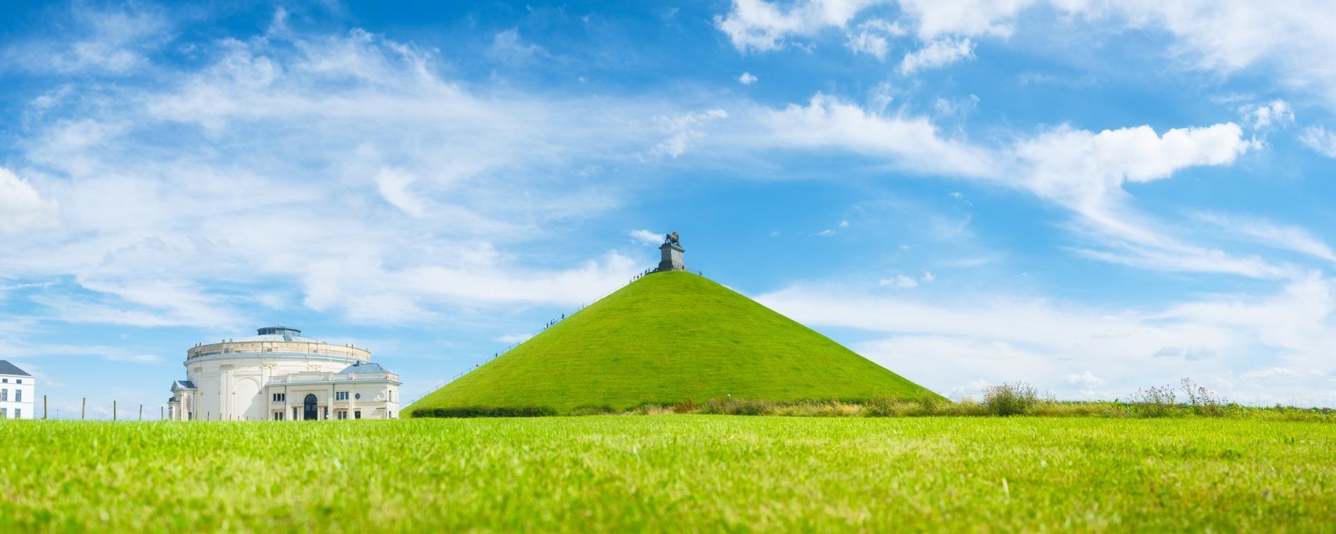 A wide shot on a summer's day with blue skies and light cloud. A hill stands in the middle of the shot in the distance with a white pavilion to the left. A vast green field sprawls across the foreground. Belgium, Waterloo.