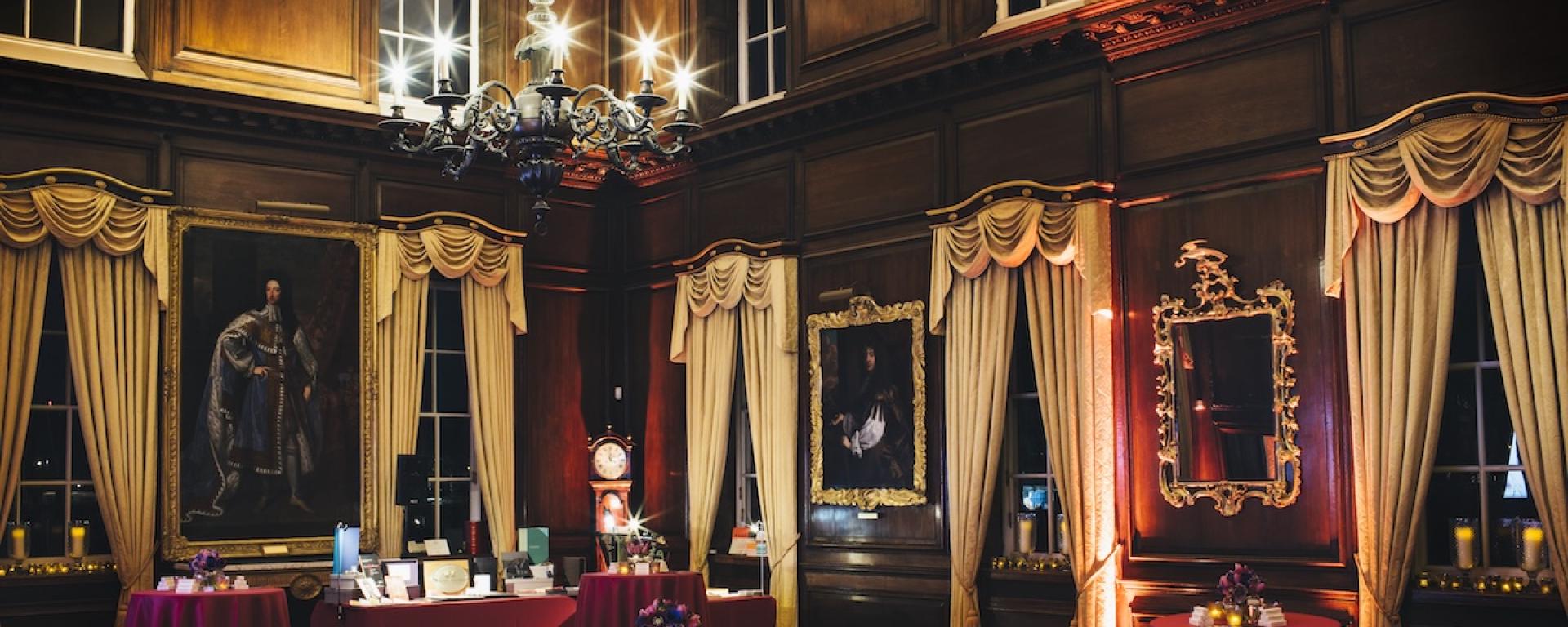 An opulent wood panelled room, with long flowing white curtains and red covered tables throughout the room. Large historic paintings hang on the walls and a warm light emphasises the night time shot.