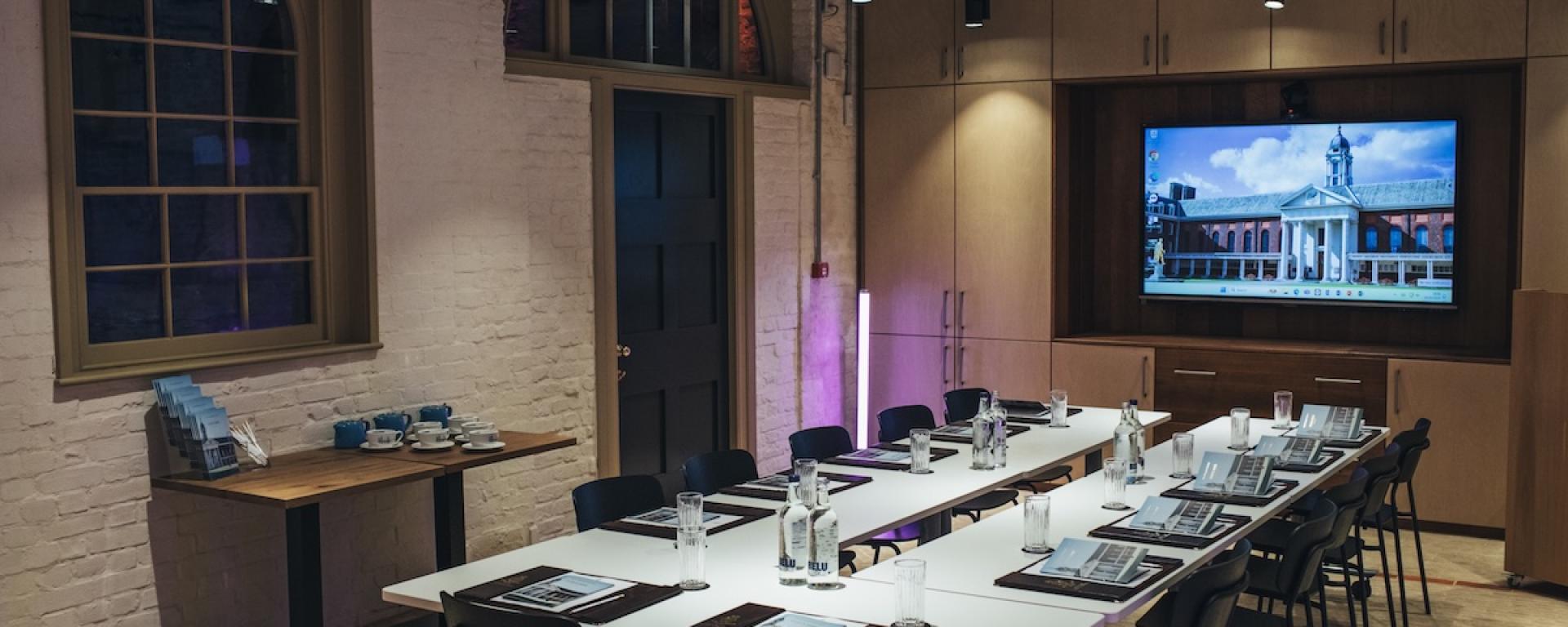 A modern meeting room with U shaped white tables set for a presentation. Each place has a glass, a notepad, and a bottle of water. Black chairs surround the tables. A large screen at the front displays an image of the Royal Hospital Chelsea. The room has exposed white brick walls, arched windows, warm overhead lighting, and built in wooden cabinetry. A side table holds stacked brochures and blue cups.