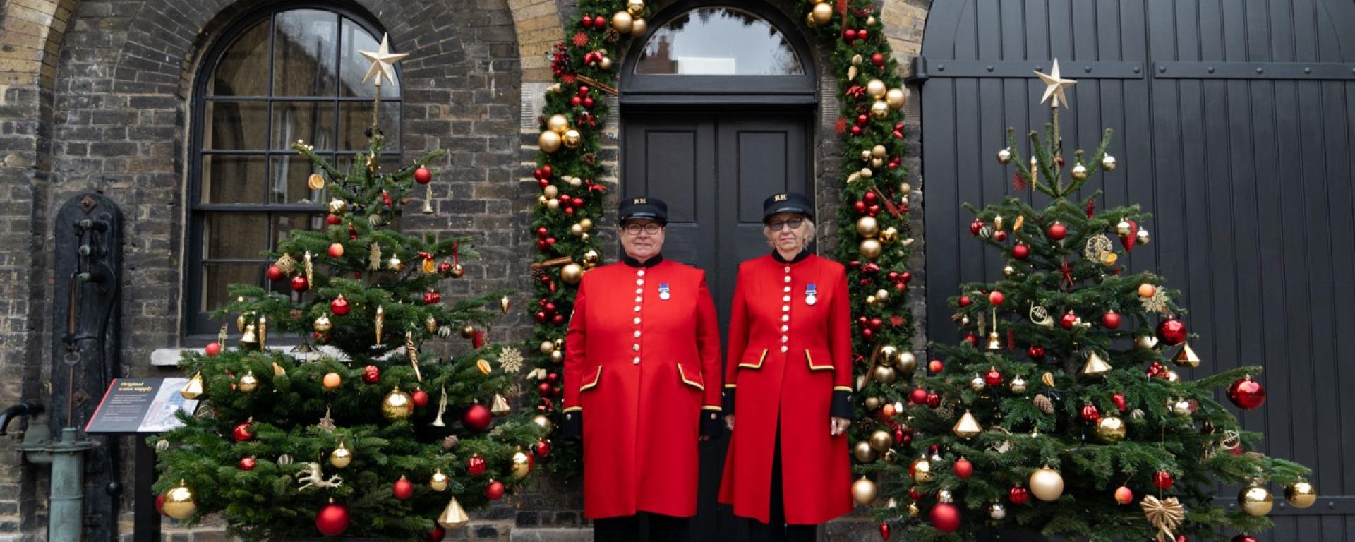 Two Chelsea Pensioners in their traditional scarlet coats and black caps stand side by side in front of a doorway decorated with a large Christmas garland. Festive Christmas trees adorned with red and gold ornaments flank them on both sides. The scene is set against the brick arches and cobblestones of the Royal Hospital Chelsea.