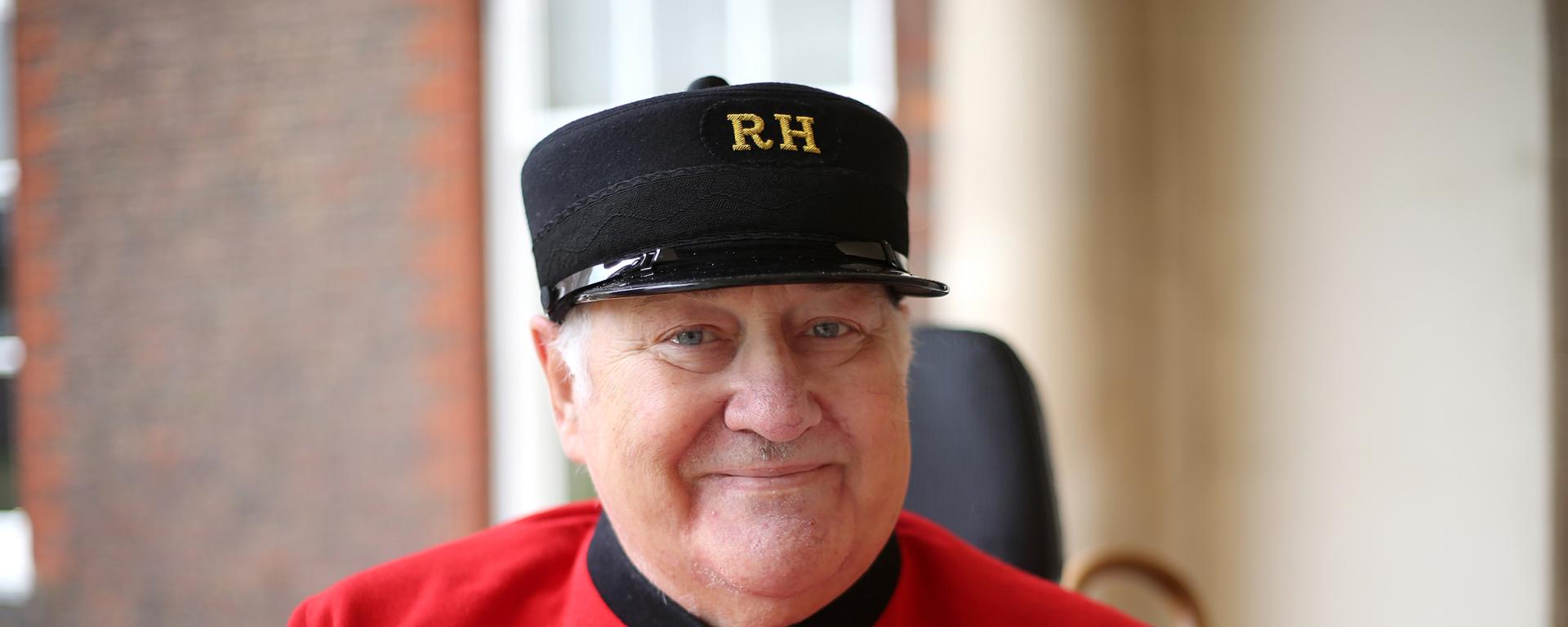 A Chelsea Pensioner wearing the traditional scarlet uniform and black cap smiles at the camera. He has several service medals displayed on his chest. The background shows the exterior of the Royal Hospital Chelsea with brickwork and white pillars softly out of focus.