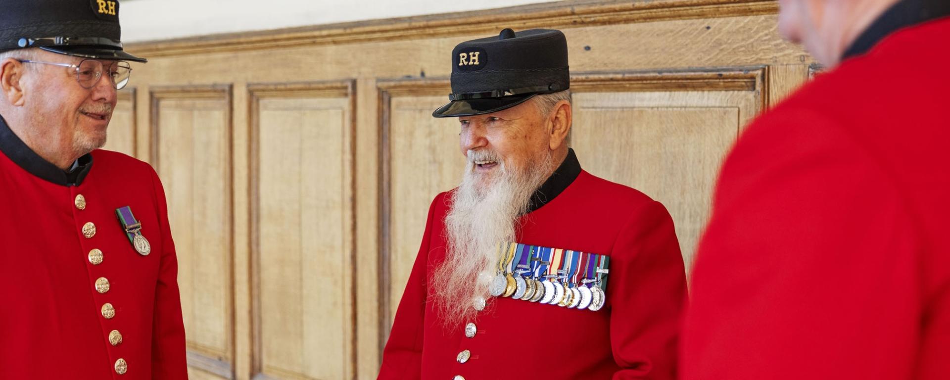 A group of Chelsea Pensioners stand in front of a wooden panelled wall, wearing their iconic scarlet uniforms with black Shako Hat emblazoned with gold "RH" lettering. The Pensioner in the centre has a long white beard and is highly decorated with almost a dozen medals.