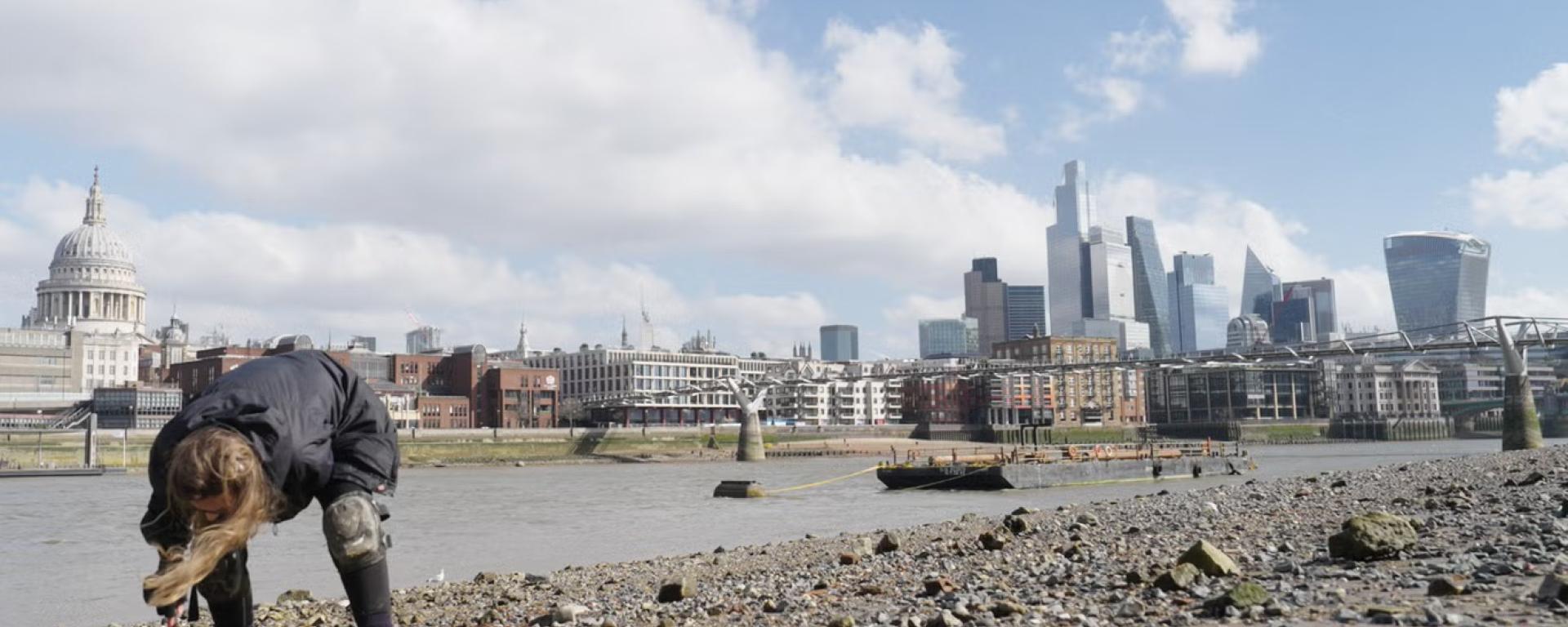 A person bends down on a rocky riverbank, examining the ground near the water’s edge. Across the river, the London skyline is visible, including St Paul’s Cathedral and modern skyscrapers. The scene is set under a bright sky with scattered clouds, contrasting natural textures with the urban cityscape.