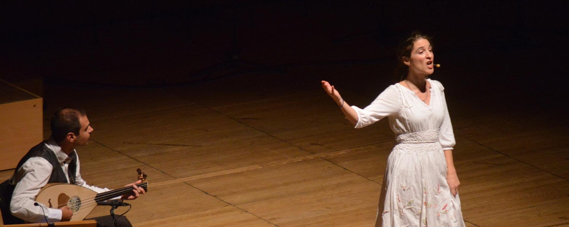 A lady stands in white dress stands on a wooden stage - one arm raised as she sings. She is basked in warm light as a well dressed man sits to the left of the stage playing a loot.