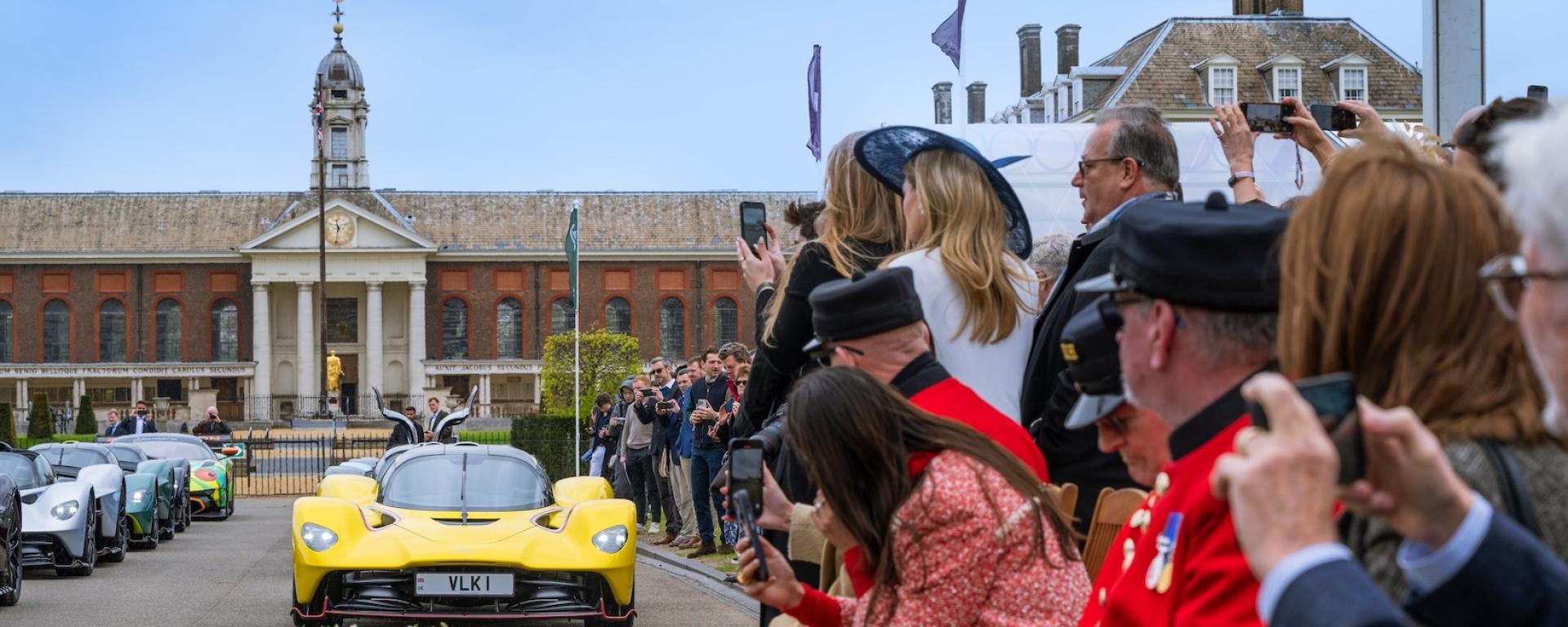 A yellow sports car stands on the south grounds of the Royal Hospital Chelsea, with the iconic building in the background and numerous spectators, including the Chelsea Pensioners dressed in their iconic scarlet uniforms, arranged alongside the right of the car.
