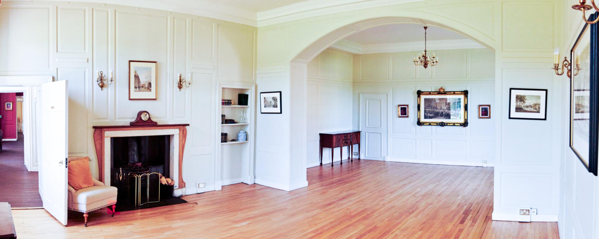 A spacious, elegant room with light wooden floors and white panelled walls, featuring classic architectural detailing and soft lighting from wall sconces and chandeliers. The room includes a fireplace with an armchair, built-in bookshelves, and framed artwork throughout. A large arched opening leads into a second, similarly styled area, creating an airy and refined atmosphere.
