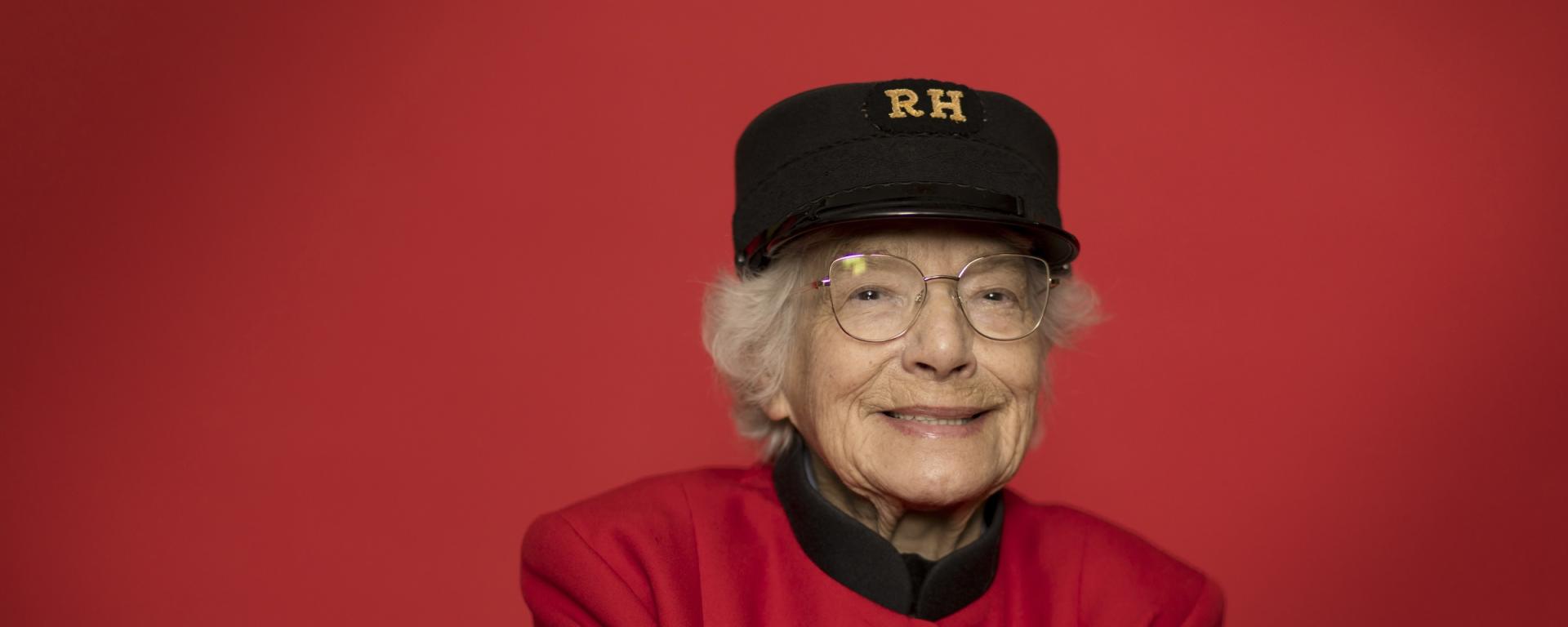 Elderly Chelsea Pensioner seated against a red backdrop, wearing a scarlet uniform with service chevrons and an RH cap, hands folded.