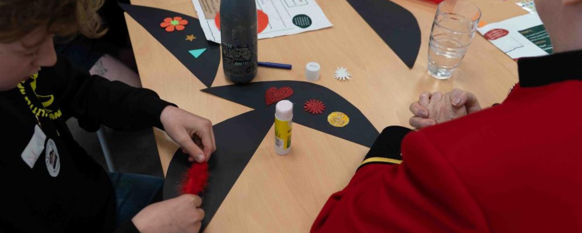 Two people sit at a table taking part in a craft activity, decorating black paper shapes with colourful stickers and materials. One person wears a red Chelsea Pensioner uniform, while the other works with a red feather and glue. Craft supplies, a water bottle, and instruction sheets are spread across the table, suggesting a shared, hands-on creative session.