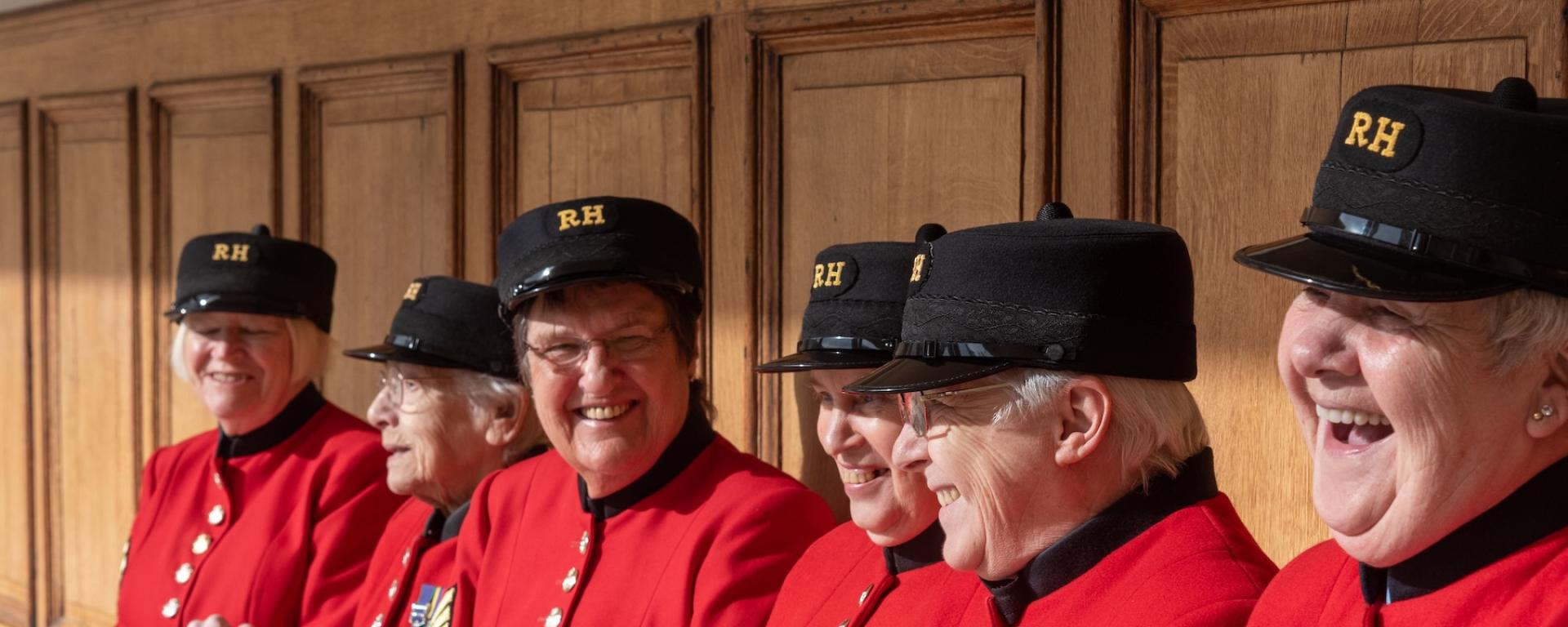 A group of Chelsea Pensioners sit side by side on a wooden bench indoors, wearing their scarlet uniforms and RH hats. They are smiling and laughing together, with medals visible on their coats. Several hold walking sticks, and the scene conveys companionship, pride, and shared humour.