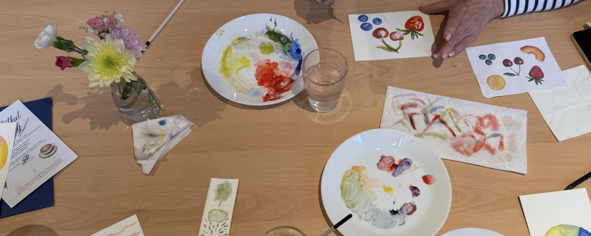 A wooden desk is littered with an array of art supplies, paints, paper plates being used at pallets and hand painted artwork.