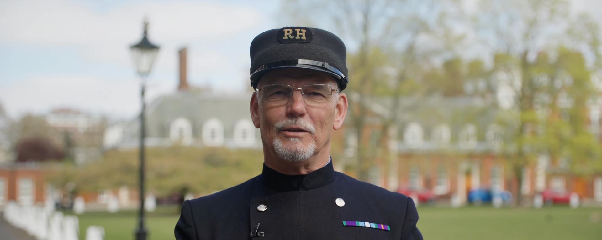 A male Chelsea Pensioners stands outside in the grounds of the Royal Hospital Chelsea wearing his "Blues" uniform and shako hat.