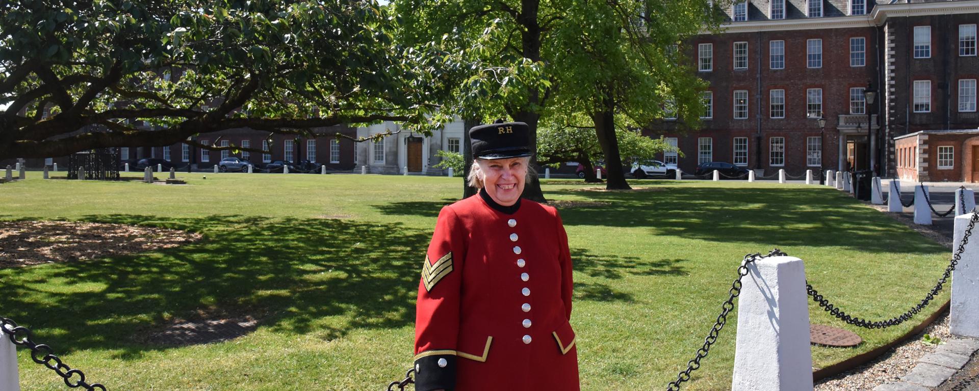 A smiling Chelsea Pensioner in a scarlet uniform stands on a sunlit lawn beneath a spreading tree, with historic buildings of the Royal Hospital Chelsea in the background.
