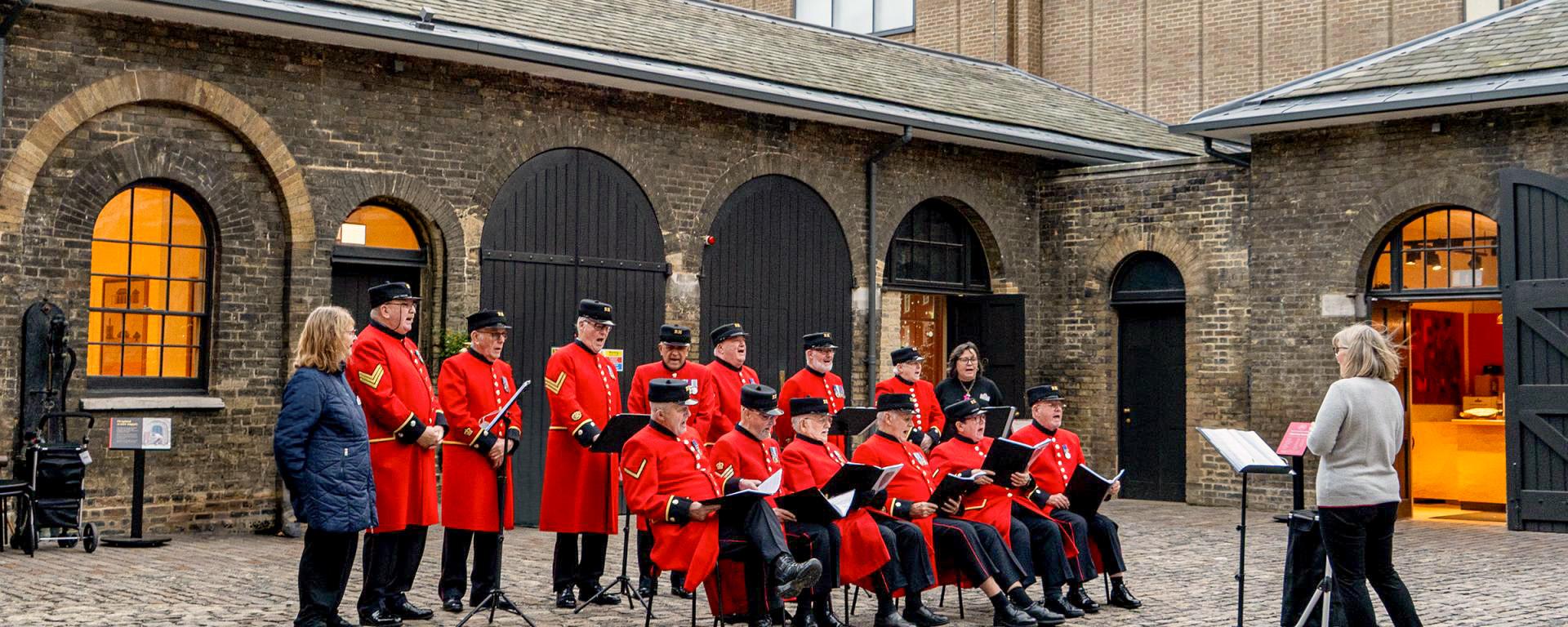 A choir of Chelsea Pensioners in scarlet uniforms sing together in the Soane Stable Yard at the Royal Hospital Chelsea, with a conductor leading them in front of the historic brick archways.