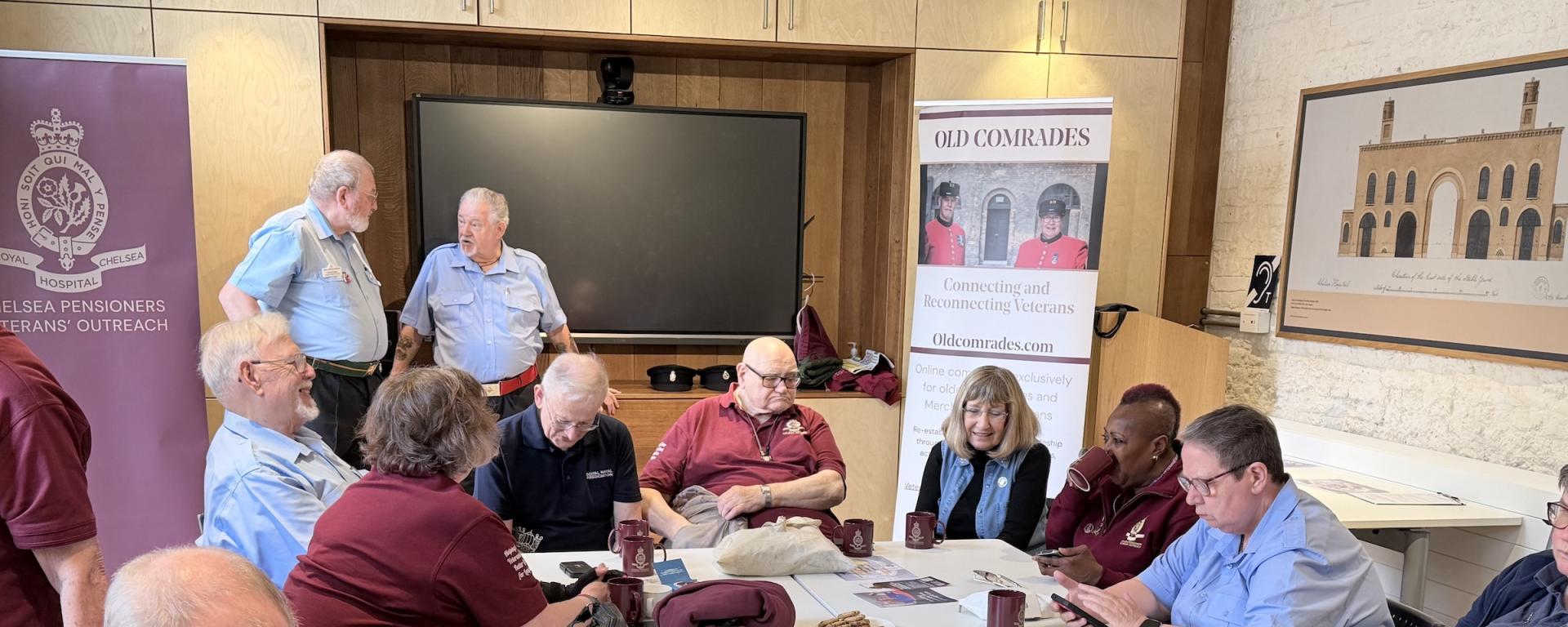 Chelsea Pensioners sit alongside veterans in a room with a large video screen and banners promoting Veterans Outreach