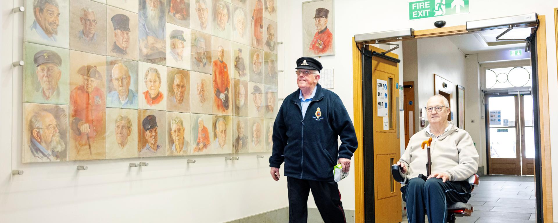 Two Chelsea Pensioners move through a bright corridor at the Royal Hospital Chelsea, passing a large wall display of painted portraits, with one man walking beside another using a mobility scooter.