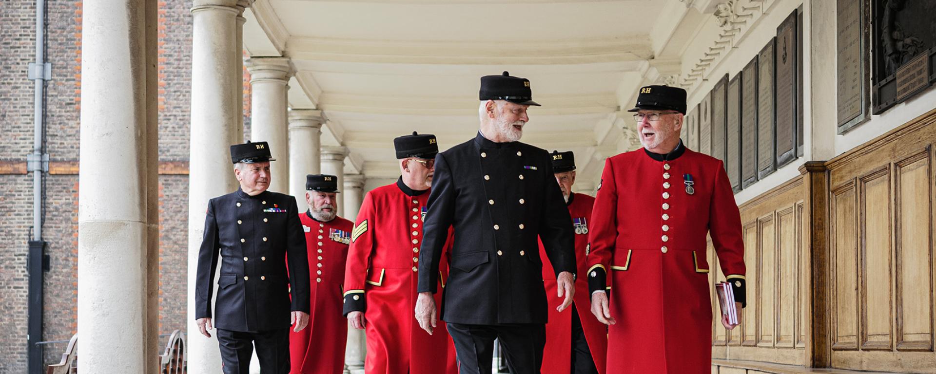 A group of Chelsea Pensioners dress in a mix of iconic scarlet and blues uniform, walk together happily along a wood panelled colonnade.