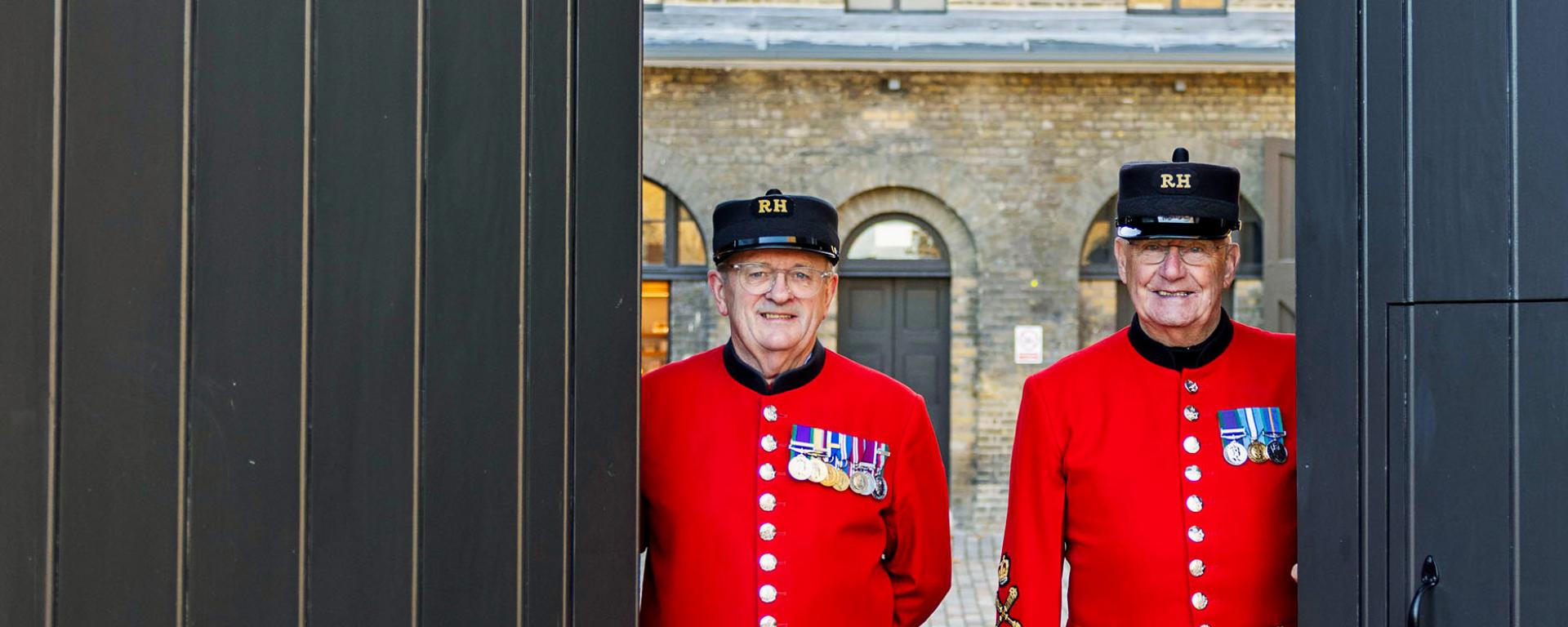Two Chelsea Pensioners in scarlet uniforms stand smiling in a doorway of the Soane Stable Yard at the Royal Hospital Chelsea, framed by large dark doors and historic brick buildings behind them.